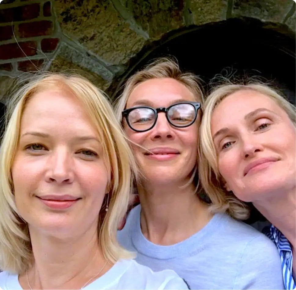 The image features three women posing closely together, with the central woman wearing black-framed glasses and a light blue t-shirt.