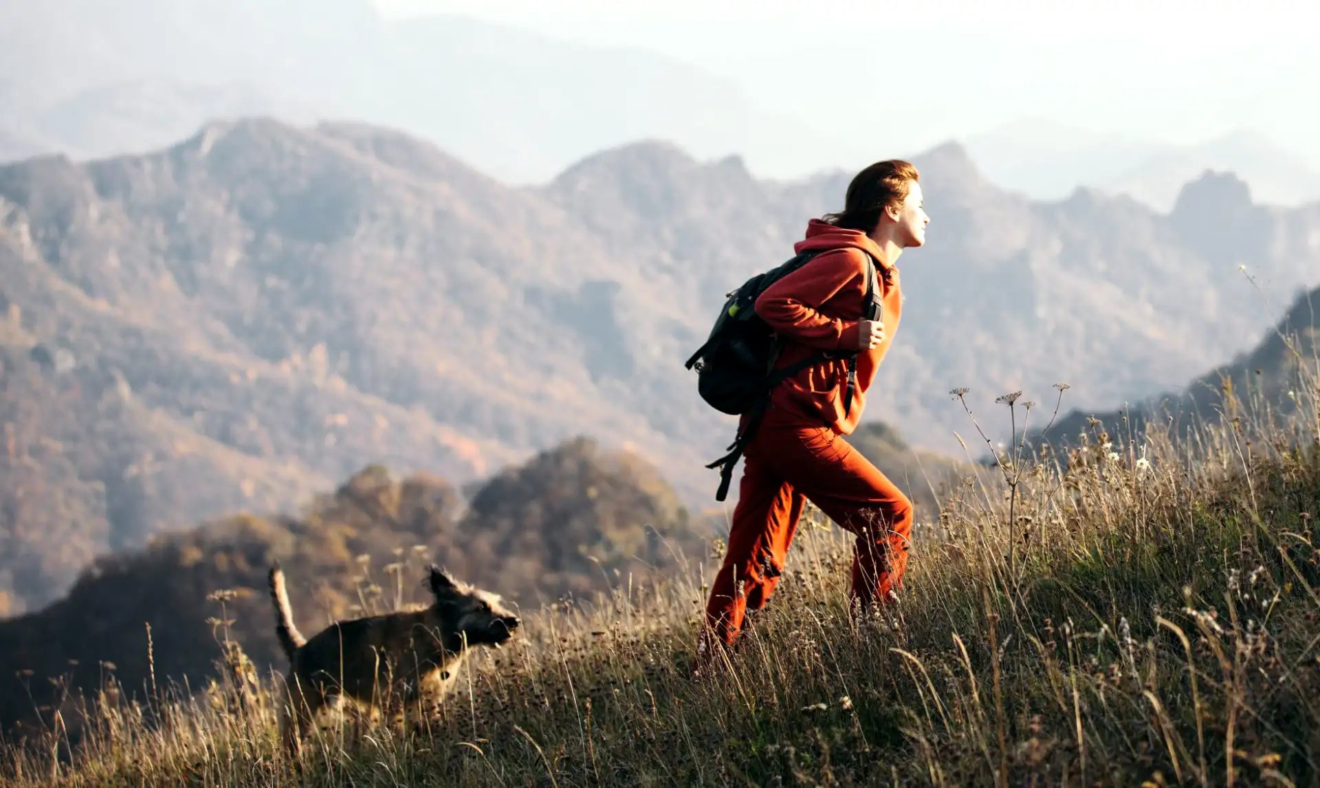 A person in a vibrant orange jumpsuit runs across a grassy hillside, carrying a black backpack.