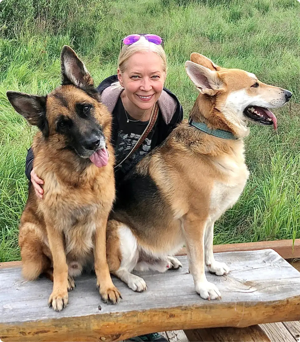A woman with blonde hair and purple sunglasses perched on her head smiles while sitting outdoors between two dogs, one a german shepherd with tan and black fur and the other a lighter-colored dog with white paws.