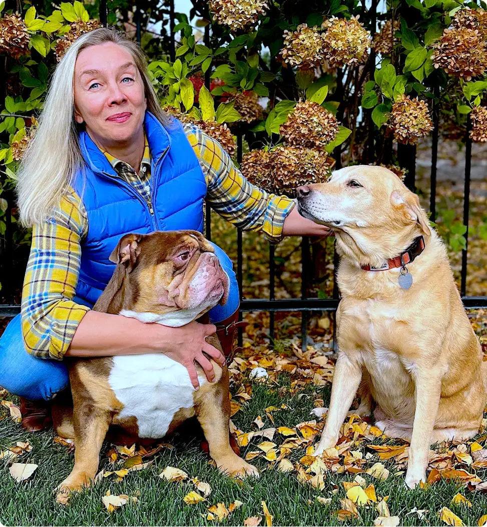 A woman with long blonde hair wearing a blue puffer vest over a yellow and green plaid shirt, crouching beside two dogs in an autumn garden.